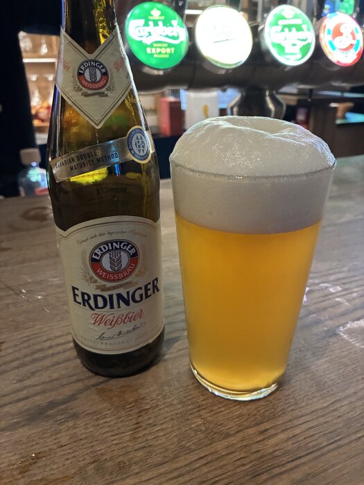 Flaska med Erdinger Weißbier och ett glas med skummande öl på en bar med flerfärgade öltappar i bakgrunden.