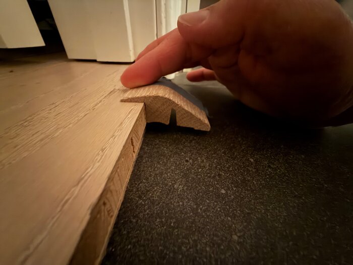 Träbit som ska användas som upphöjd tröskel, placeras mellan golv och dörrkarm. En hand visar bitens form och passform, fokus på stöd och fäste.