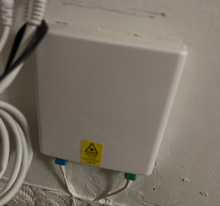 Vit fiberbox monterad på vägg med kablar kopplade, varningstext synlig.