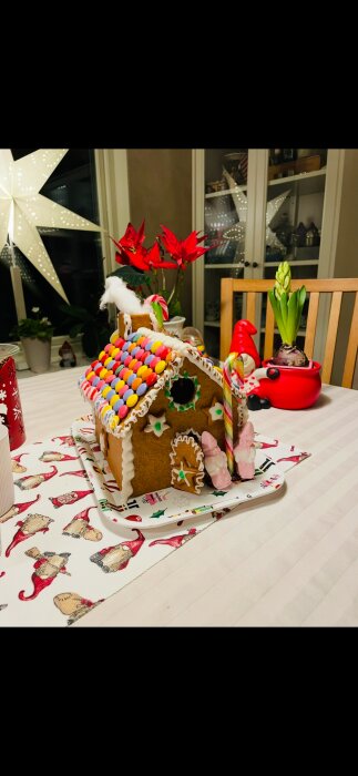 Pepparkakshus dekorerat med färgglada godisar och vit glasyr på ett bord med juldukar, röda blommor och ljusstake i bakgrunden.