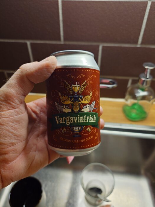 Hand håller burk med etiketten "Vargavintrish 2012", Sälen Fjällbryggeri, framför en brun kaklad vägg i ett kök.