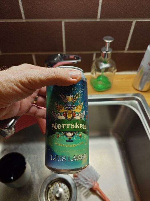 Hand håller i en burk öl med etiketten "Norrsken Ljus Lager" från Sälen bryggeri framför ett kökshandfat.