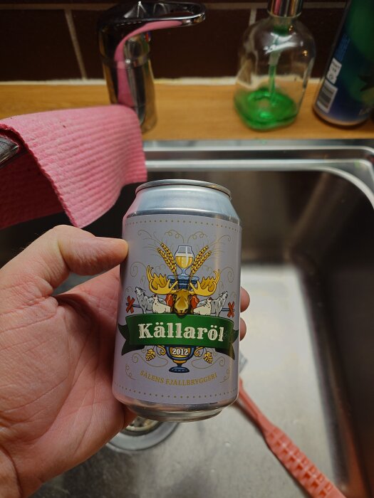 En hand håller en ölburk med etikett märkt "Källaröl" från "Sälens Fjällbryggeri" över en diskbänk, med diskborste och diskho i bakgrunden.