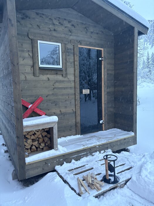 Bastu i snölandskap med vedstapel, pall och vedklyv vid dörren, redo för kvällseldning.