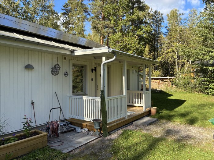 Vit farstukvist med träpanel och räcken på ett hus, omgivet av grönska och en solig trädgård.