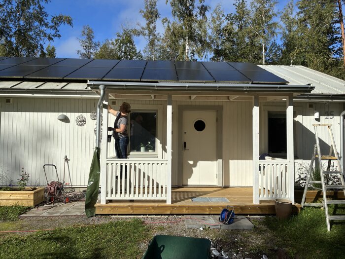 Person arbetar på nybyggd farstukvist vid vit villa med solpaneler, omgiven av träd och trädgårdsutrustning, en stege står bredvid.