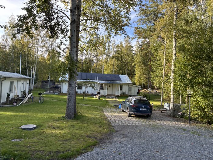 Hus med nybyggd farstukvist, solpaneler på taket, cykel vid sidan och bil parkerad på uppfarten, omgivet av träd och grönska.