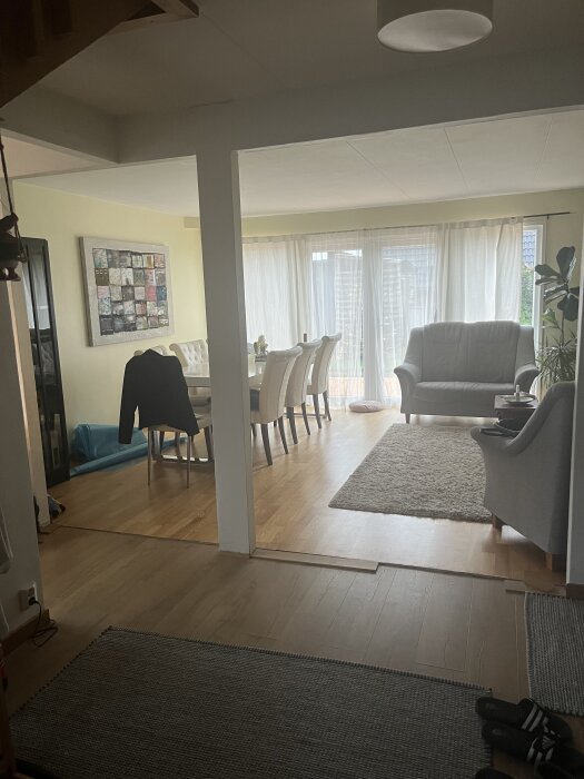 Interiör av ett vardagsrum med en bärande vägg och mittstolpe, matbord med stolar och ljusinsläpp från stora fönster med gardiner.