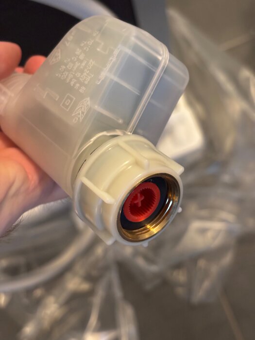 VVS-komponent i plast med mässingsgängor och röd insats, hålls i hand mot suddig bakgrund.