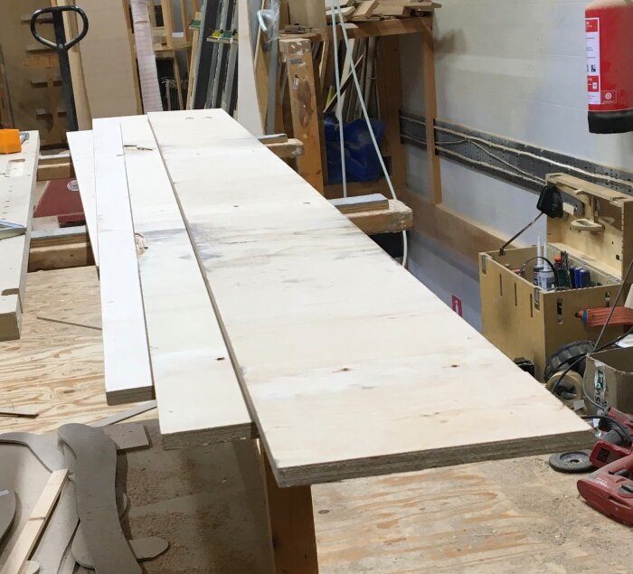 Tunna plywoodskivor ligger på ett arbetsbord i en snickarverkstad, omgivna av verktyg och byggmaterial.