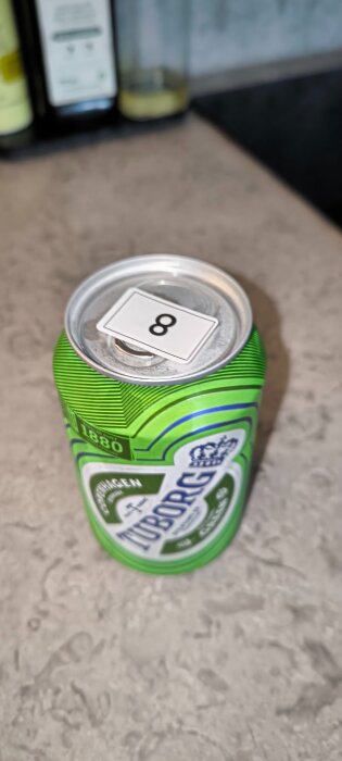 Grön ölburk på grå bänkskiva med ett klistermärke med siffran "8" på toppen. I bakgrunden syns två suddiga flaskor.