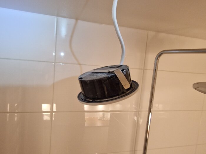 En inbyggd svart taklampa med en vit sladd hänger nedåt i ett rum med kakelväggar. En metallclips hålls mot lampan.