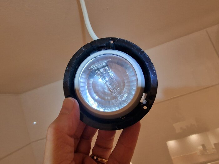 Hand håller en takmonterad spotlight med en glödlampa; ringen närmast glaset är avlägsnad för att ge tillgång till lampan.