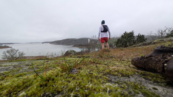 Person i träningskläder tittar ut över en sjö från en mossig klippa. Mulen himmel och skogsklädda kullar i bakgrunden.