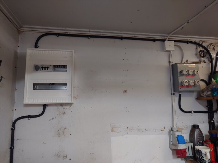 Vit vägg i garage med elcentral, kablar och uttag installerade, synliga fläckar och hylla med verktyg.