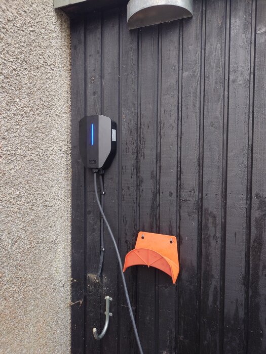 Laddstation för elbil monterad på en svart trävägg med ett orange kabelhållare och metallkrok.