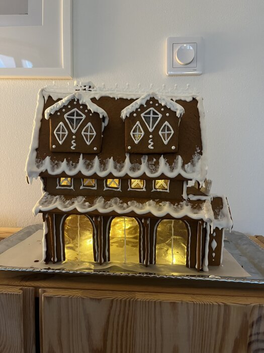 Pepparkakshus med två våningar, dekorerat med kristyr, står på en bricka. Fönstren är upplysta, vilket ger en varm och inbjudande känsla.