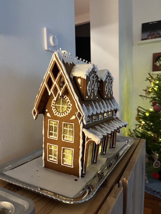 Pepparkakshus med glasyrdetaljer och belysning, placerat på en bricka, med en julgran synlig i bakgrunden.