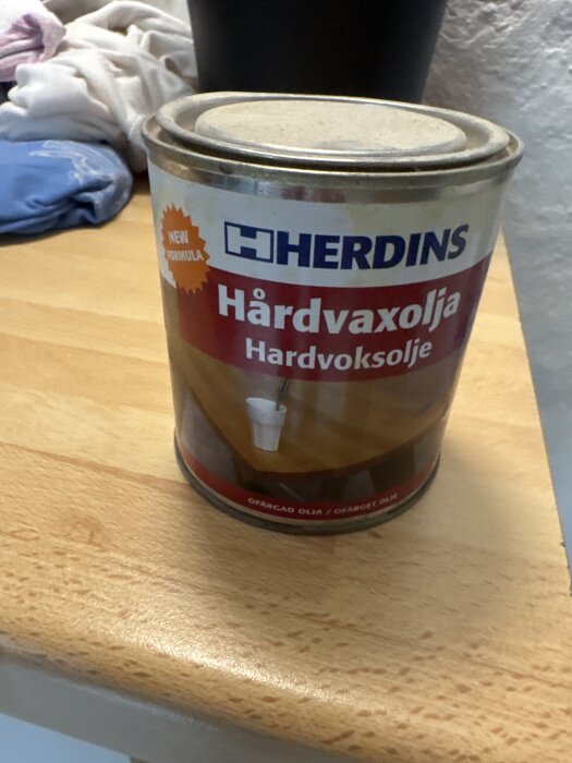 Burk med Herdins hårdvaxolja på en träyta, omgiven av kläder.