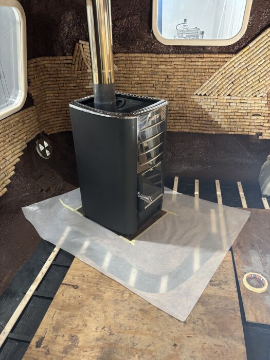 Kamin placerad på trägolv med kartongmall under för att utforma en träram som följer de rundade hörnen mot väggens korktextur.