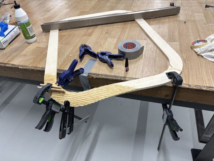 På bilden syns en träbåge fixerad med klämmor och tejp på ett bord. Verktyg och limflaskor ligger bredvid.