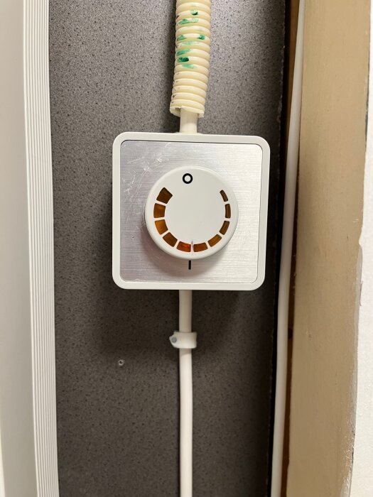 Rund dimmer inkopplad i väggen, styr en fläkt. Beige sladd med grön märkning leder till dimmern. Bakgrund av grå väggpanel.