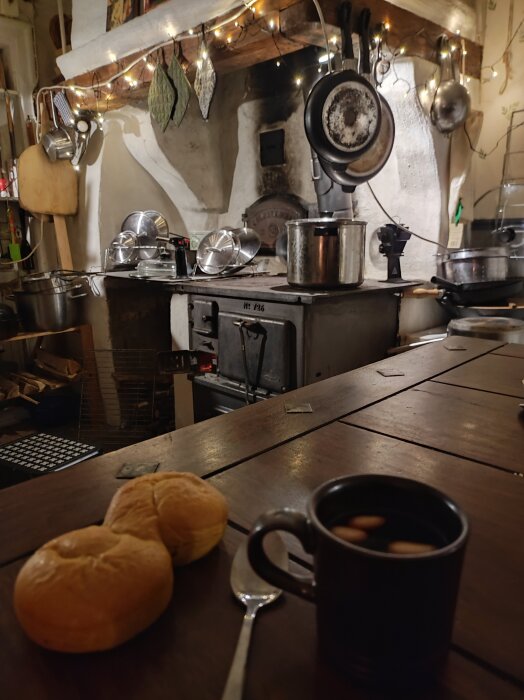 Vedspis med grytor och kastruller, ljusslinga ovanför. En kopp glögg med mandlar och två bullar på bordet.