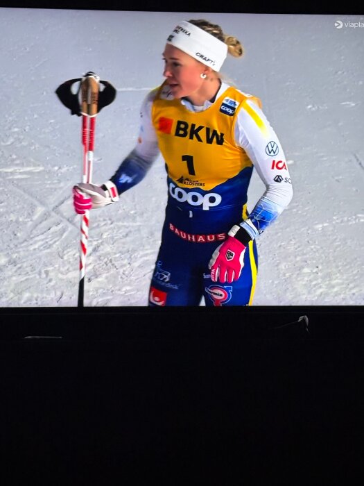 Skidåkare i gul väst och blå dräkt på snö, hållande skidstavar, med fokus på sportaktivitet.