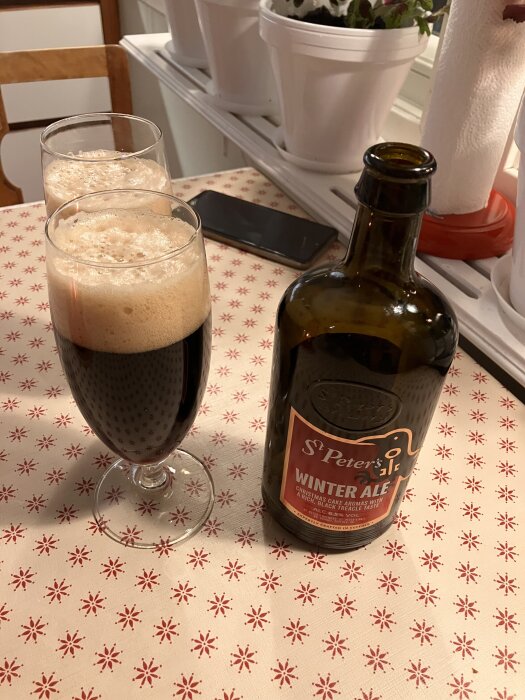 Två glas med mörk öl och skum bredvid en öppnad flaska St. Peter's Winter Ale på ett bord med rött och vitt mönster.