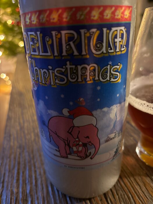 Flaska med etikett "Delirium Christmas" och en rosa elefant med tomteluva, bredvid ett glas med mörk Belgisk Ale på bord vid julgran.