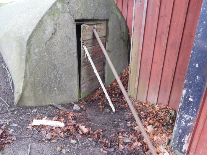 Gammalt träskydd med stöttande träpinne, delvis dolt bakom ett rödmålat plank, omgiven av löv och jord.