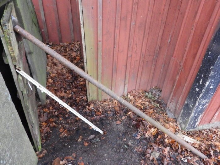 Rostig stång som stöttar en gammal träport, omgiven av löv vid en rödmålad trävägg.