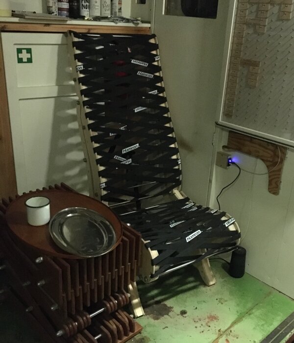 Stol med svart bandnät och en trästruktur står bredvid ett bord av vikta lameller med en mugg och bricka på, i ett rum med verktygstavla.