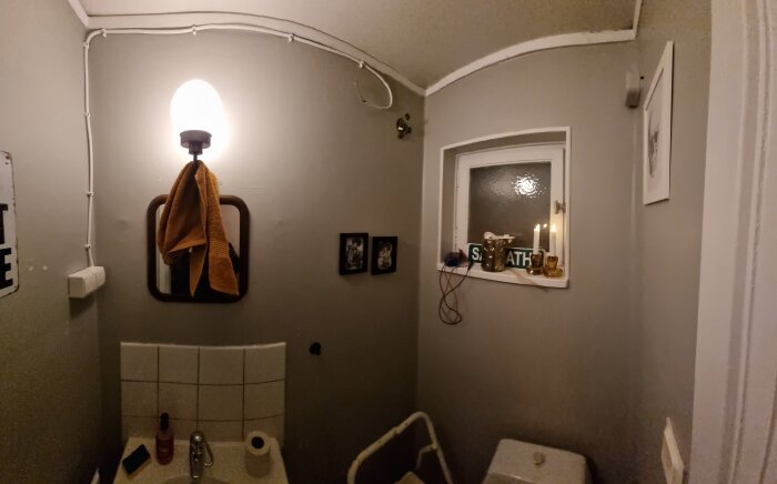 Badrum med lampa ovan spegel och synliga kablar vid tak. Infälld kopplingsdosa på väggen. Flera fotografier och föremål på fönsterbrädan.