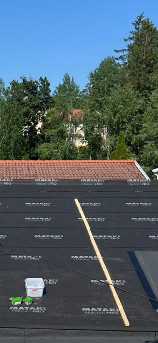 Tak täckt med Mataki Haloten PRO diffusionöppen duk, träregel och en plastlåda med verktyg, samt omgivande träd och tegelpannetak i bakgrunden.
