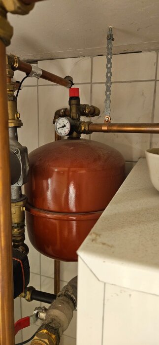 Rött expansionskärl med manometer under ett rörsystem, placerat i ett rum med kaklade väggar.
