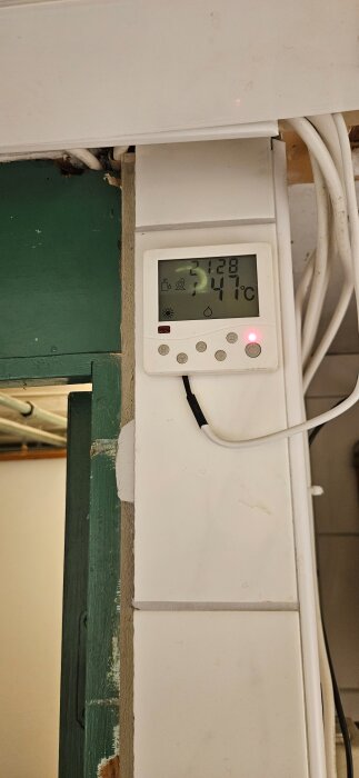 Väggmonterad termostat med digital display som visar aktuell temperatur och tid, omgiven av sladdar och vit inredning.