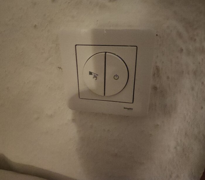 Väggmonterad dimmer med två knappar, en för justering av ljusstyrka och en för av/på, installerad på ojämn beige vägg.