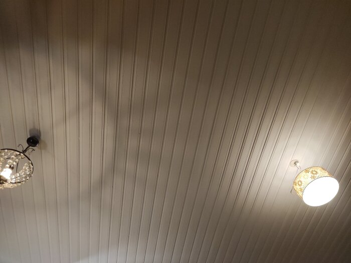Två taklampor tända i ett rum, en med en kristallkrona och den andra med ett mönstrat lampskärm, monterade på ett vitmålat trätak.