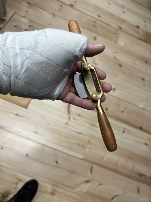 En hand i bandage håller en ny, gyllene hyvel med trähandtag över ett ljust trägolv.