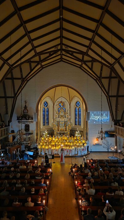Kyrka interiör med ett luciatåg framför altaret, omgivna av åskådare och levande ljus, under ett traditionellt firande.