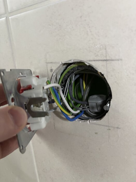 Elinstallation med vit vägguttag, exponerade kablar i en rund väggdosa. En hand håller uttaget vid sidan av dosan.