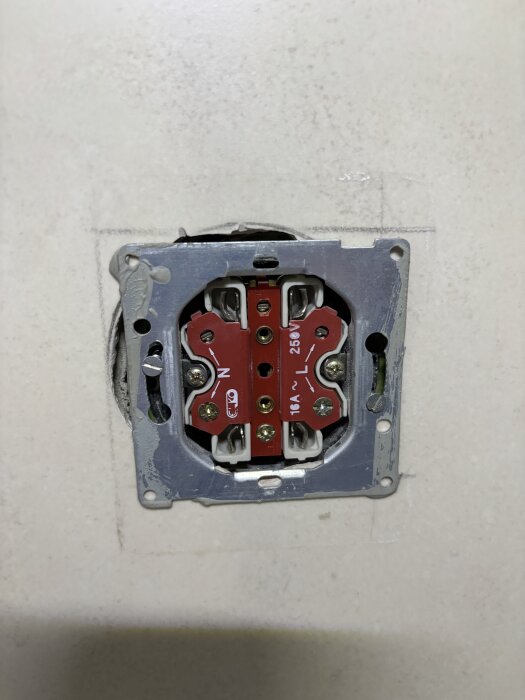Elektrisk väggkontakt utan täckplatta, visande kopplingspunkter och ledningar, monterad på en vit vägg.