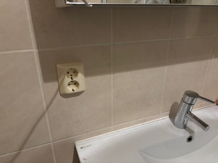 Eluttag med två uttag sitter på kaklad vägg ovanför ett handfat med en kromad kran i ett badrum.