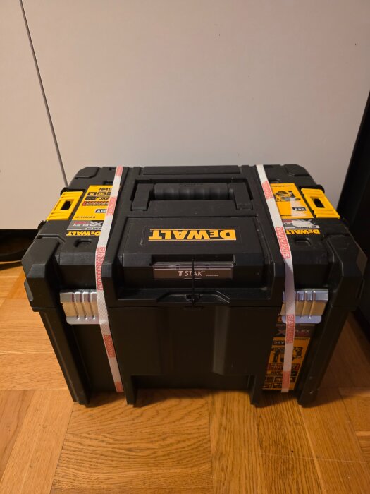 En svart Dewalt verktygslåda med TSTAK märke, stående på ett trägolv, omgiven av vita och gula klistermärken.
