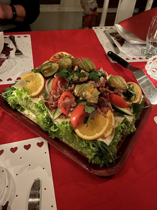 Smörgåstårta på ett rött dukat bord, dekorerad med kyckling, bacon, frukt och grönsaker.
