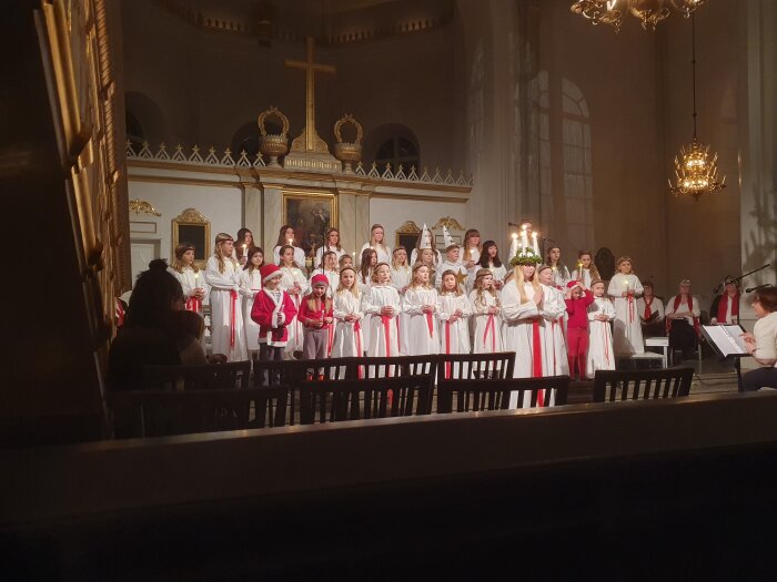 En grupp barn i vita kläder och röda band uppträder i en kyrka. En flicka med Luciakrona står i mitten. Publikum och dirigent syns delvis.