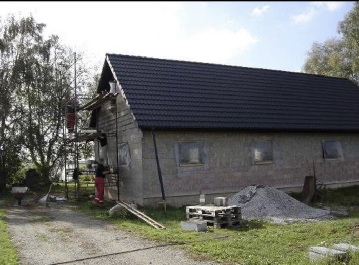Hus med svart tak och ställning på sidan, halvgjort med byggmaterial framför, placerad i en grönskande miljö.