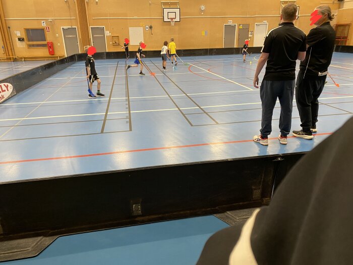 Barn spelar innebandy i en idrottshall medan två vuxna i sportkläder står och tittar på.