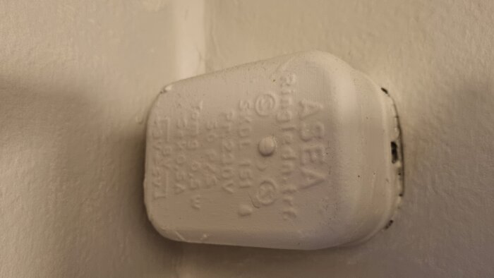 En vit dosa monterad på väggen med text och symboler, kan vara en gammal kopplingsdosa eller en del av en ringklocka.